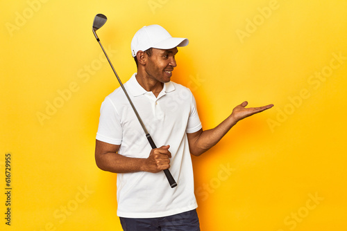 African American man golfer  yellow studio backdrop  showing a copy space on a palm and holding another hand on waist.