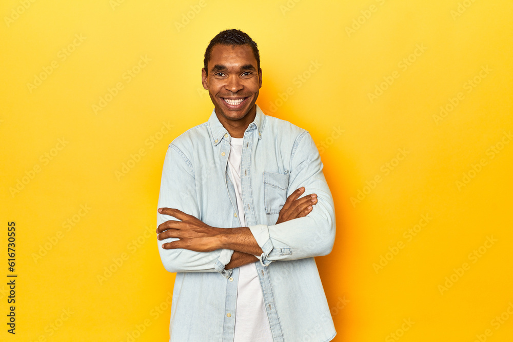 African American man in blue shirt, yellow studio, who feels confident, crossing arms with determination. African American man in blue shirt, yellow studio, who feels confident, crossing arms with determination.