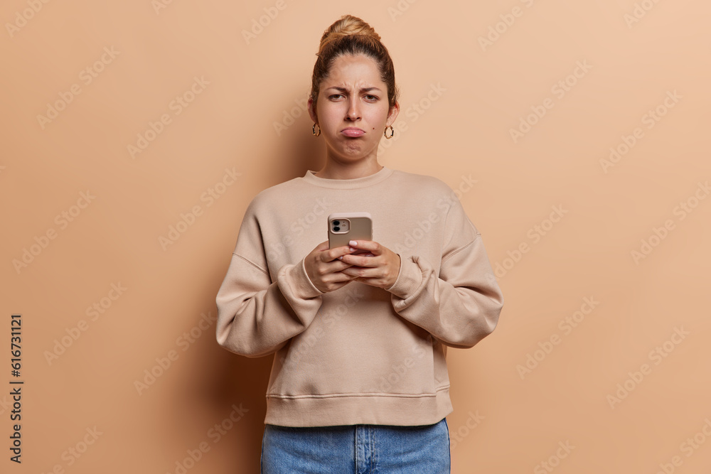 Displeased young woman with hair bun holds mobile phone has offended expression as reads bad news dressed in casual pullover and jeans isolated over brown background. Negative human emotions Displeased young woman with hair bun holds mobile phone has offended expression as reads bad news dressed in casual pullover and jeans isolated over brown background. Negative human emotions