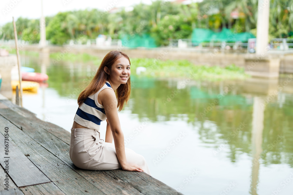 Asian cute teenage girl with long hair Sit and relax in your free time on the pier. By the river on weekends with a beaming face. A rural canal with clear water