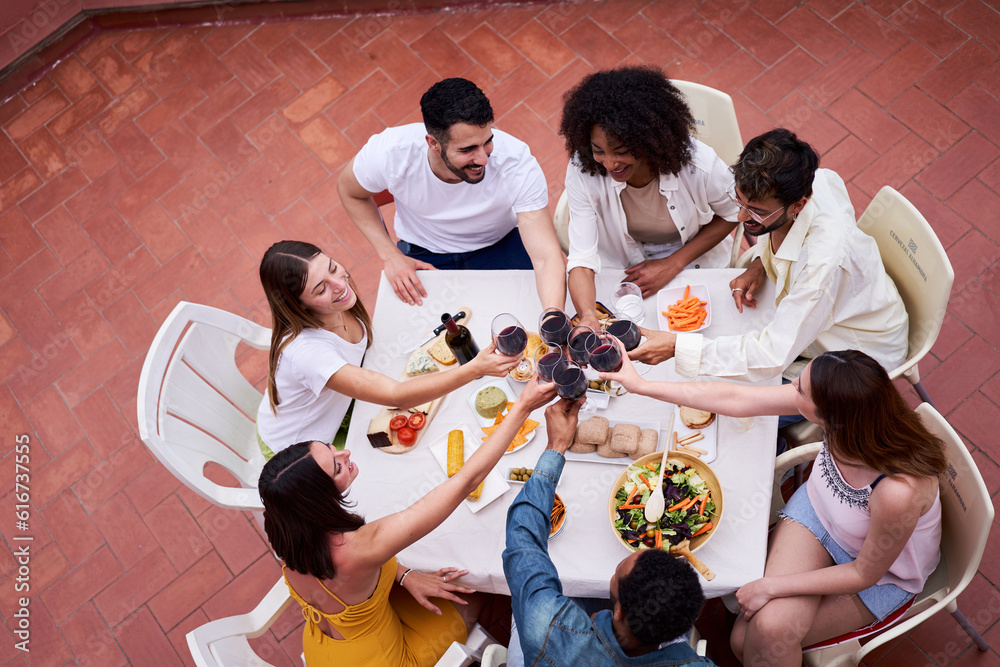 High angle view of people toasting. Happy group of Multiracial friends having dinner outside in a terrace celebrating with red wine. High angle view of people toasting. Happy group of Multiracial friends having dinner outside in a terrace celebrating with red wine.