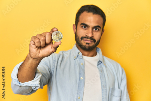 Young Latino man showcasing a physical Bitcoin coin, yellow studio background.