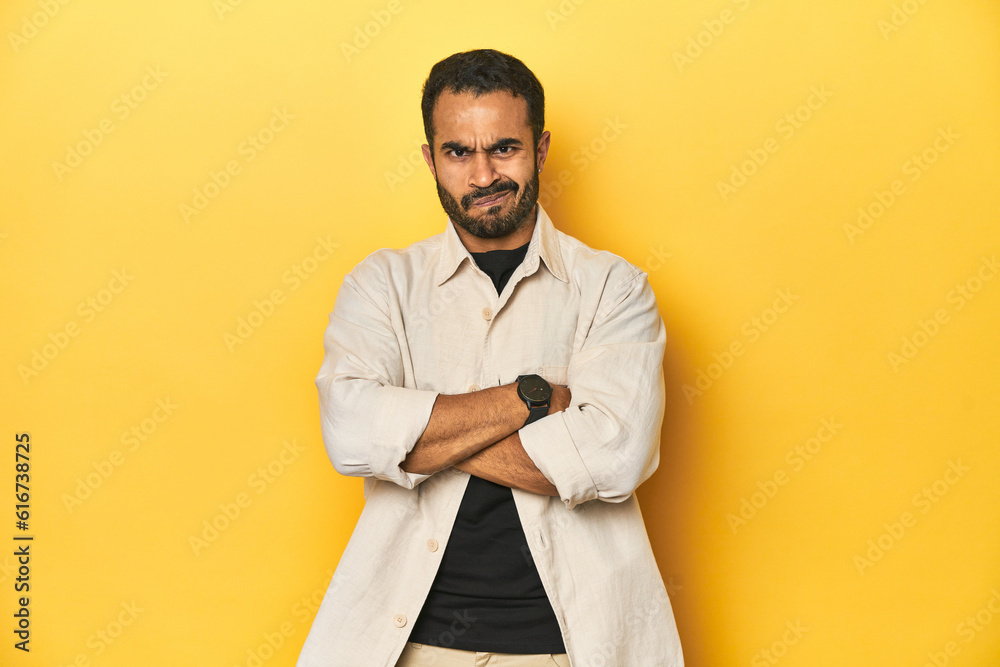 Obraz premium Casual young Latino man against a vibrant yellow studio background, frowning face in displeasure, keeps arms folded.