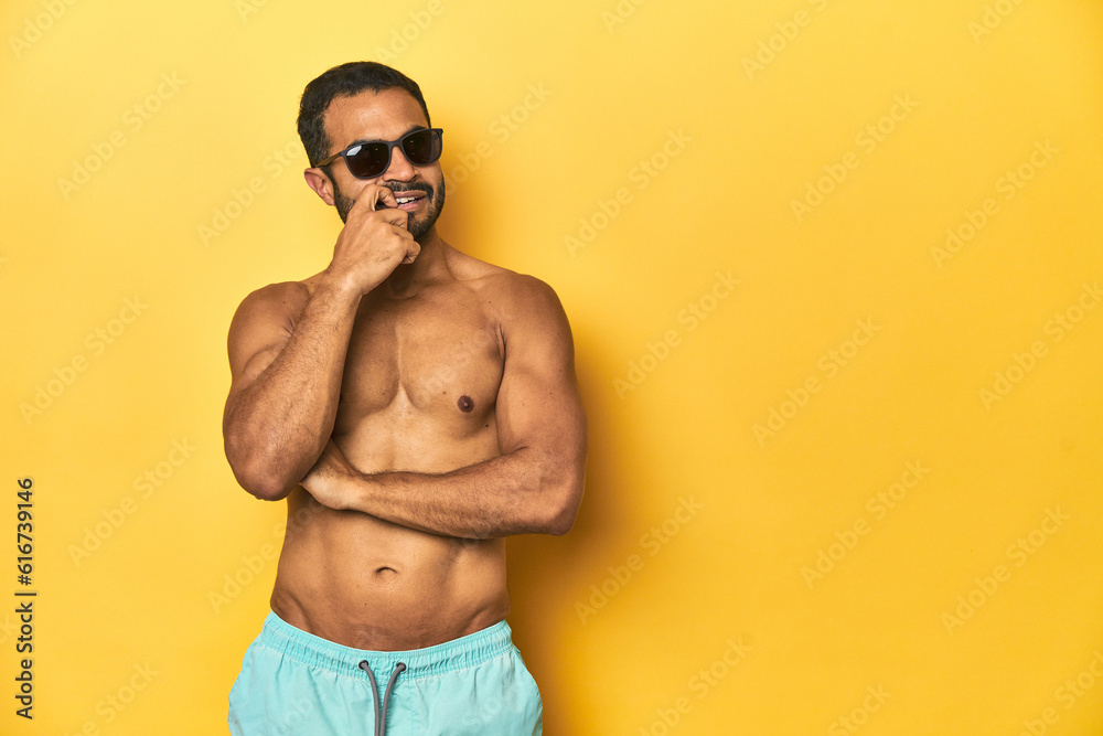Fit young Latino man in swimwear and sunglasses, yellow studio background, relaxed thinking about something looking at a copy space. Fit young Latino man in swimwear and sunglasses, yellow studio background, relaxed thinking about something looking at a copy space.