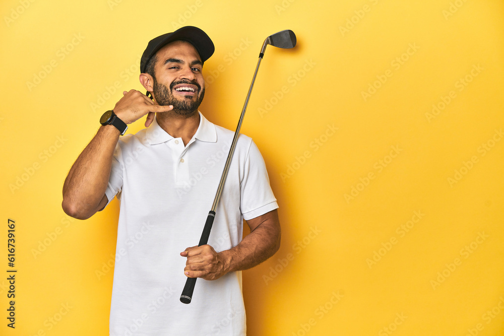 Young Latino golfer with club and cap on a yellow studio background, showing a mobile phone call gesture with fingers.