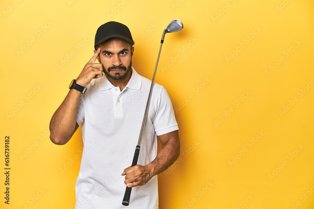 Young Latino golfer with club and cap on a yellow studio background, pointing temple with finger, thinking, focused on a task. Young Latino golfer with club and cap on a yellow studio background, pointing temple with finger, thinking, focused on a task.