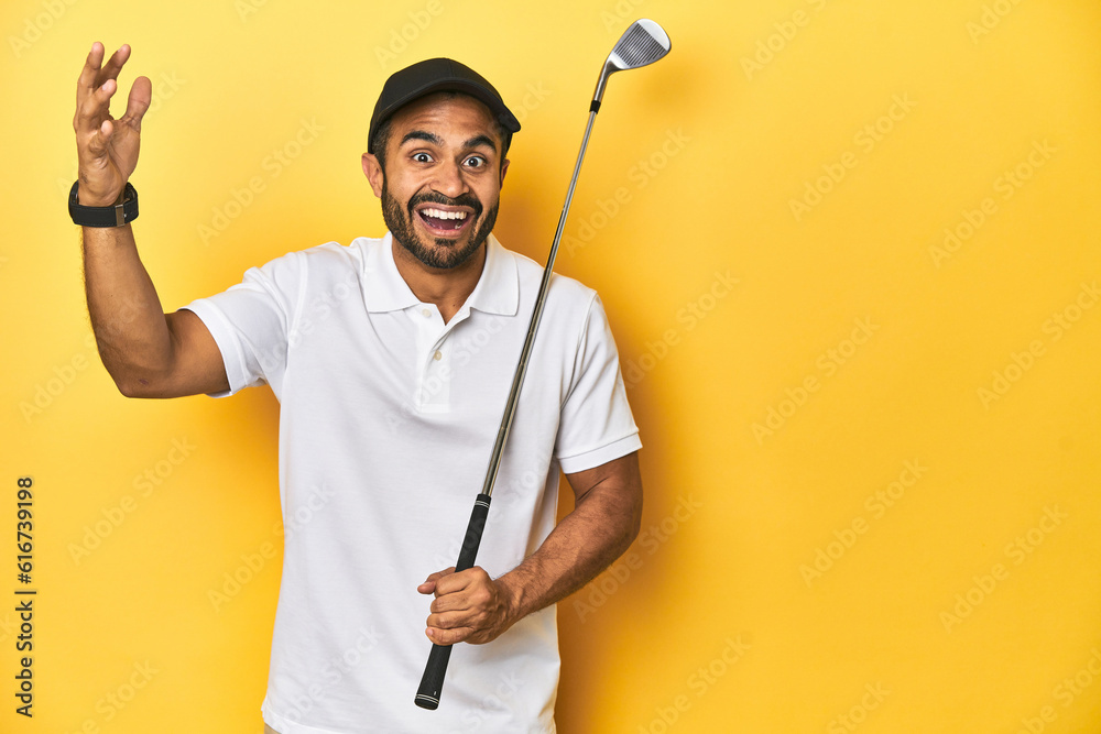 Young Latino golfer with club and cap on a yellow studio background, receiving a pleasant surprise, excited and raising hands.