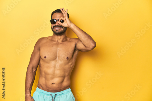 Fit young Latino man in swimwear and sunglasses, yellow studio background, excited keeping ok gesture on eye.