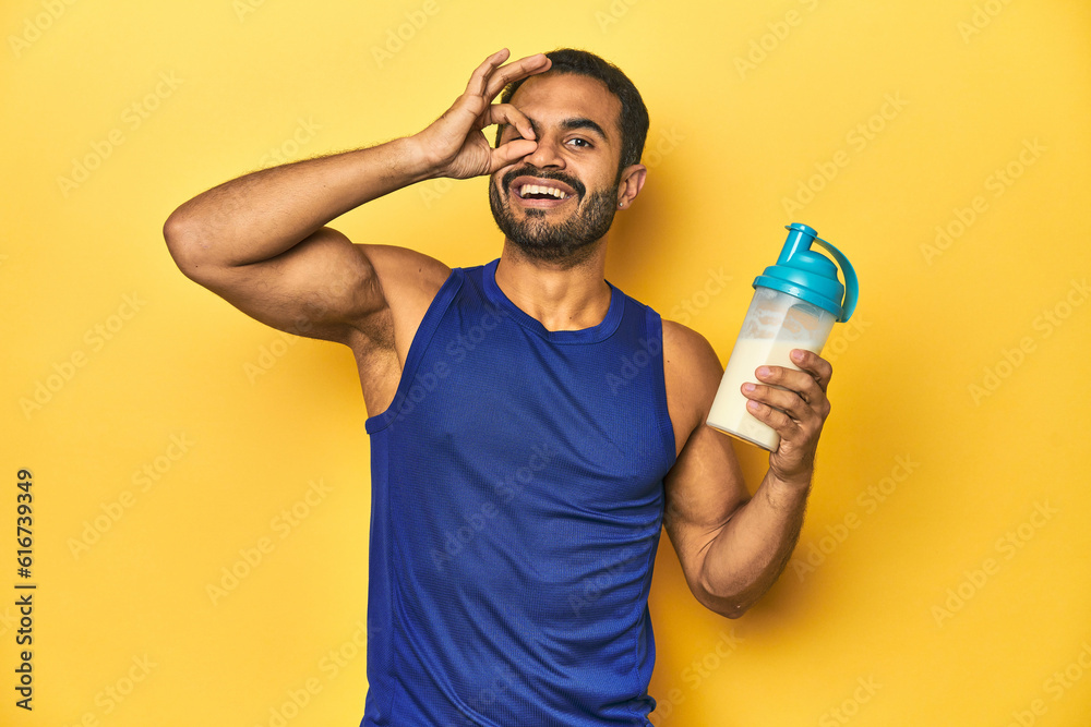 Athletic young Latino man holding a protein shake against a yellow background excited keeping ok gesture on eye. Athletic young Latino man holding a protein shake against a yellow background excited keeping ok gesture on eye.