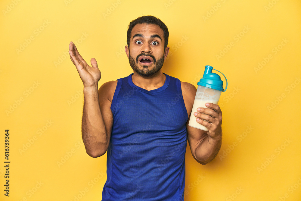 Athletic young Latino man holding a protein shake against a yellow background surprised and shocked. Athletic young Latino man holding a protein shake against a yellow background surprised and shocked.