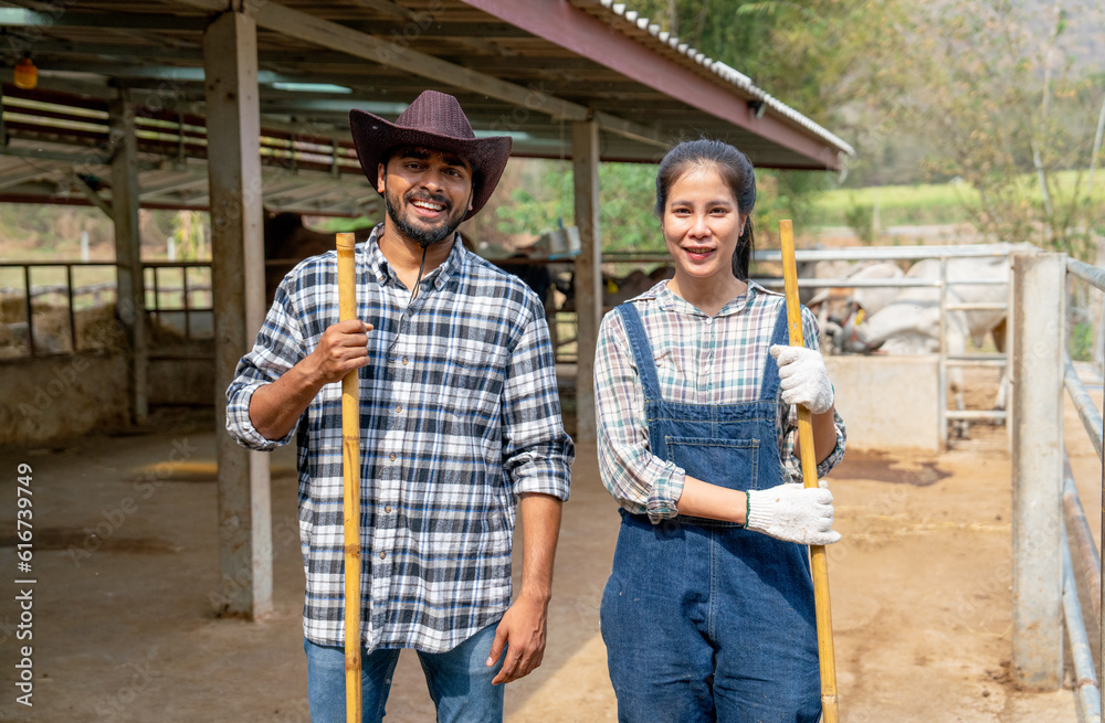 Portrait of Asian man and woman farmers carry broom and look at camera with smiling also stand in front of stable of cow look relax after clean the area.