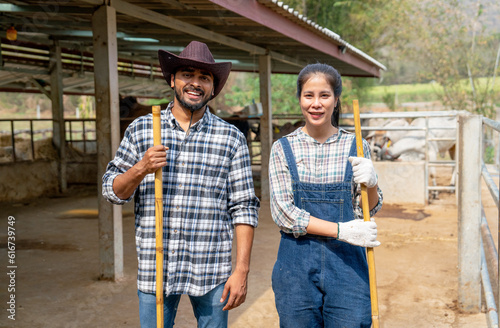 Portrait of Asian man and woman farmers carry broom and look at camera with smiling also stand in front of stable of cow look relax after clean the area.