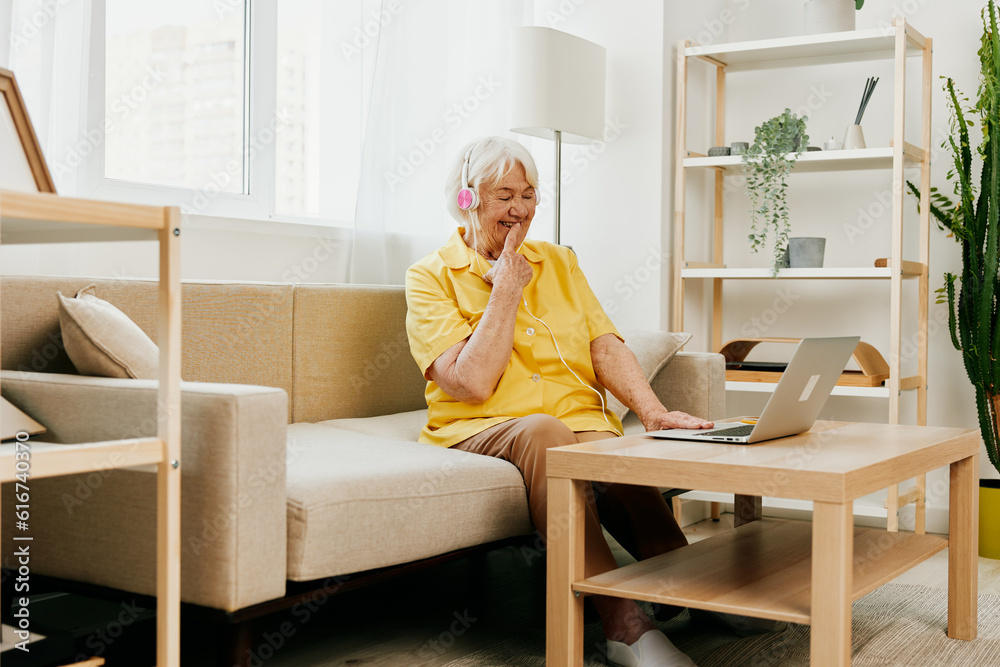 An elderly woman wearing headphones with a laptop sitting on the couch at home and working in a yellow shirt in front of a window, surprise, the lifestyle of a pensioner. An elderly woman wearing headphones with a laptop sitting on the couch at home and working in a yellow shirt in front of a window, surprise, the lifestyle of a pensioner.