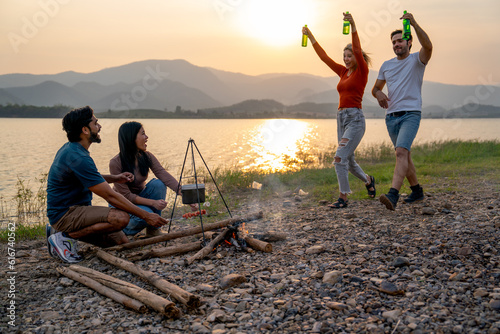 Caucasian man and woman bring bottle of drinking and walk to join with their friends during party or camping near lake of public park with sunset.