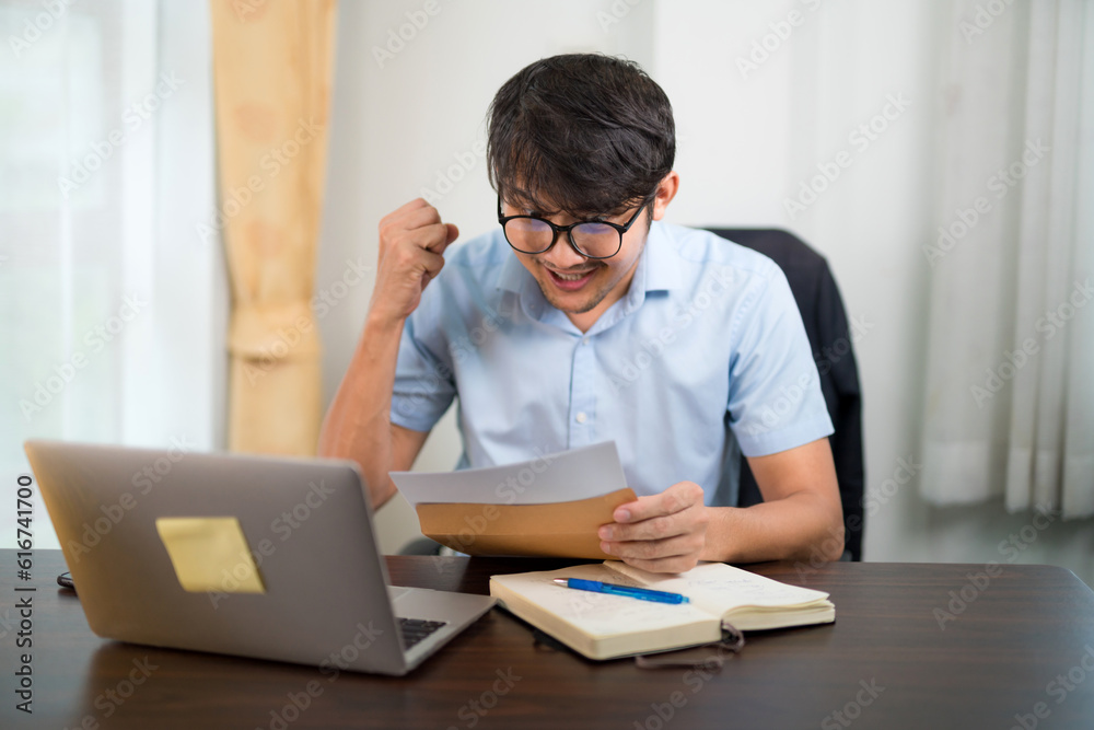 A handsome young man in glasses showing surprised expression while reading acceptance letter with laptop on desk at home. A handsome young man in glasses showing surprised expression while reading acceptance letter with laptop on desk at home.