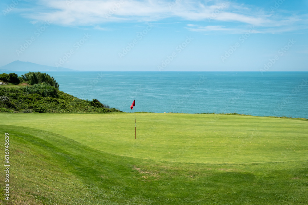 Campo de golfe com uma bandeira vermelha a meio e o mar ao fundo num dia ensolarado Campo de golfe com uma bandeira vermelha a meio e o mar ao fundo num dia ensolarado