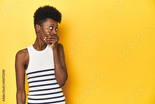 Vibrant African woman  white tank-top  yellow studio  thoughtful looking to a copy space covering mouth with hand.
