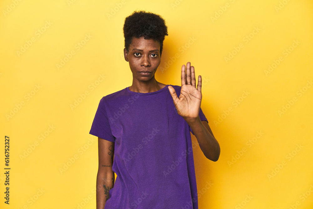 African woman in violet tee, yellow studio scene, standing with outstretched hand showing stop sign, preventing you. African woman in violet tee, yellow studio scene, standing with outstretched hand showing stop sign, preventing you.