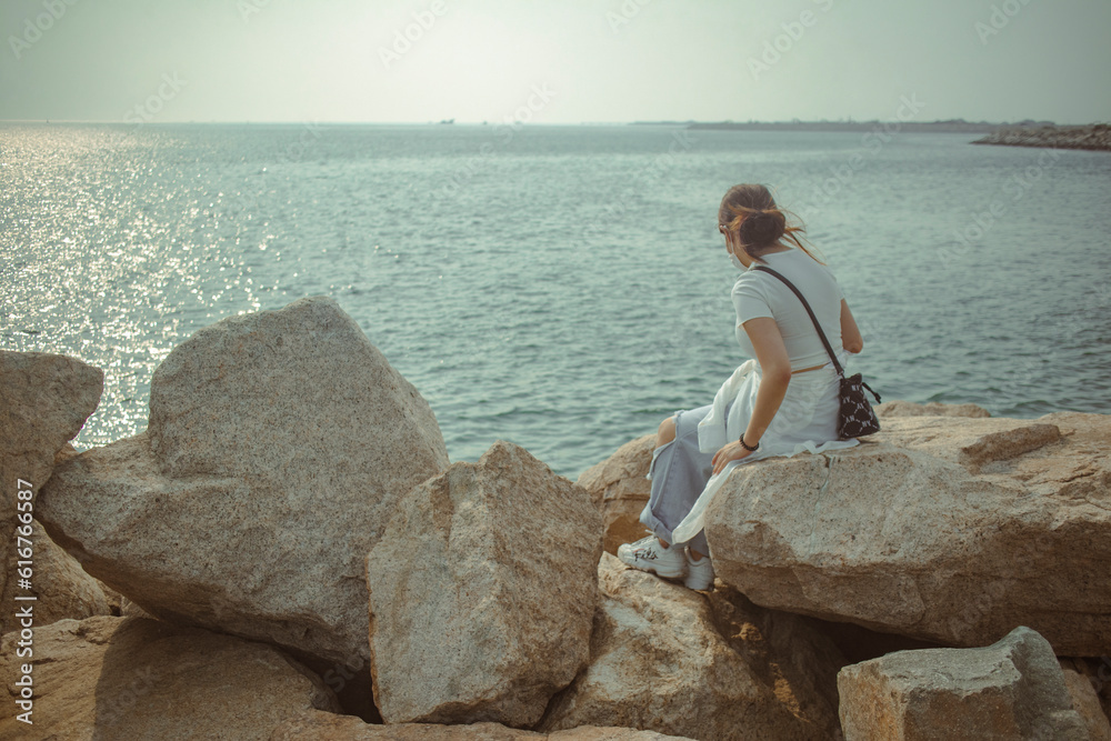 A girl is watching the golden sea on a giant rock by the coastline