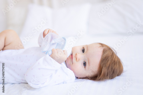 a baby girl of six months on a bed in white clothes sucks a bottle of water or milk, a small child on a cotton bed at home woke up in the morning eating, baby food concept