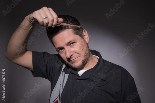 Horizontal studio portrait of a 40 year old man in rockabilly style combing his toupee with a steel folding butterfly comb
