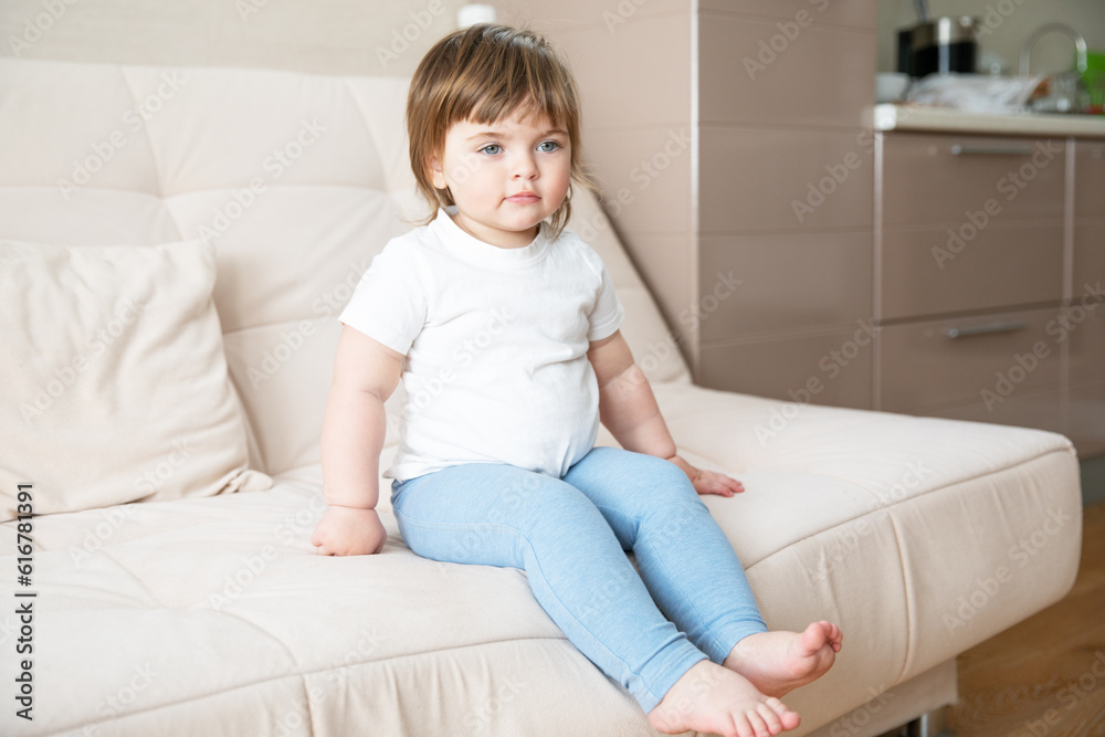 Beautiful little Caucasian girl in white T-shirt and blue pants sitting on light sofa in room. Beautiful little Caucasian girl in white T-shirt and blue pants sitting on light sofa in room.