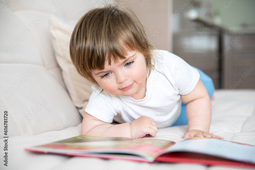 Caucasian little girl of two years with blue eyes in a white T-shirt lies on a sofa with a book. Caucasian little girl of two years with blue eyes in a white T-shirt lies on a sofa with a book.