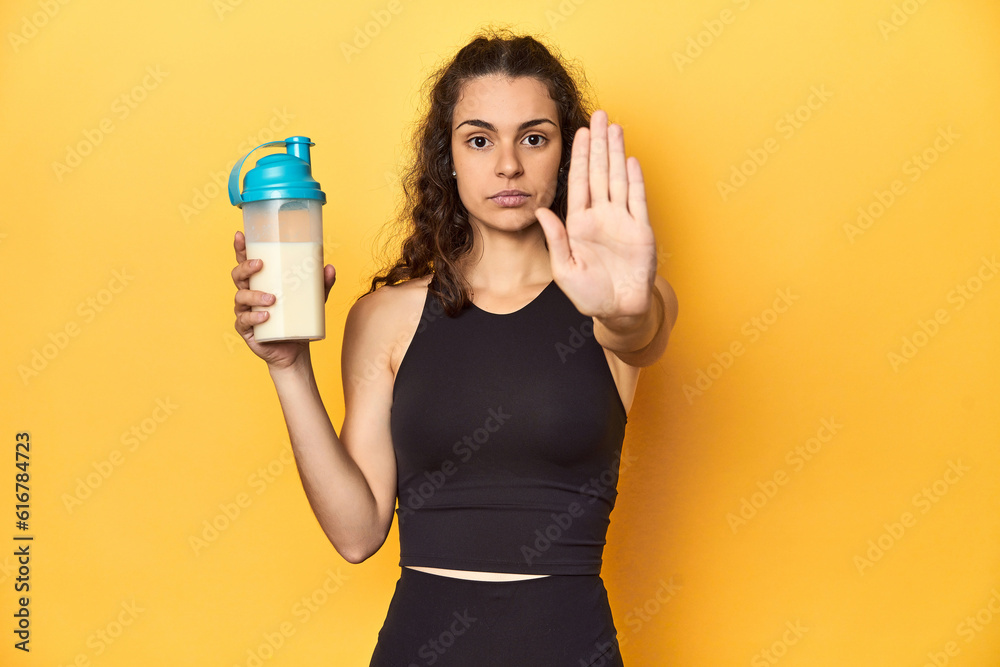 Woman holding a protein shake, in sportswear, standing with outstretched hand showing stop sign, preventing you.