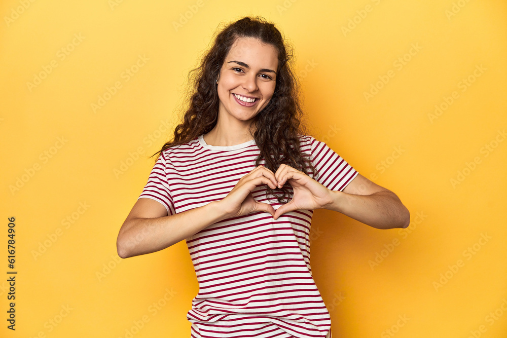 Young Caucasian woman, yellow studio background, smiling and showing a heart shape with hands. Young Caucasian woman, yellow studio background, smiling and showing a heart shape with hands.
