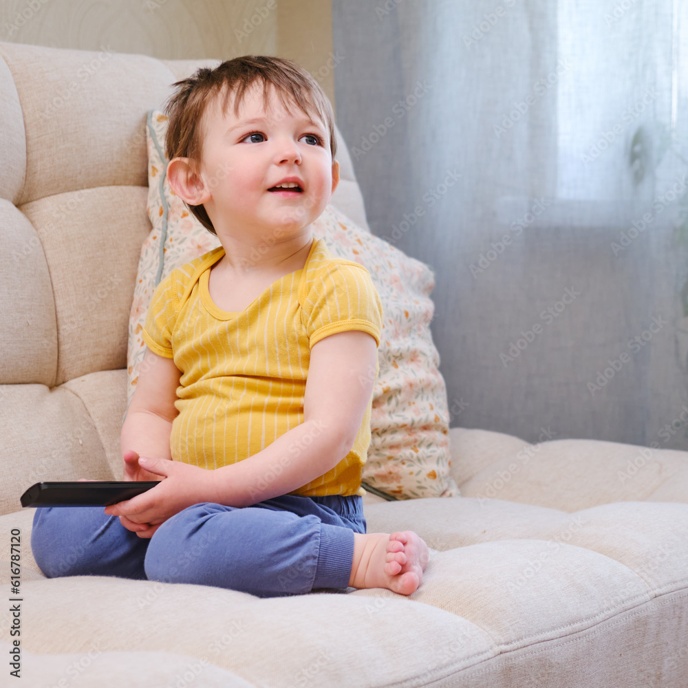 Toddler watches their favorite cartoon on TV while sitting comfortably on the sofa. The smiling child watches cartoons on TV, sitting on the beige sofa. Kid aged about two years (one year ten months) Toddler watches their favorite cartoon on TV while sitting comfortably on the sofa. The smiling child watches cartoons on TV, sitting on the beige sofa. Kid aged about two years (one year ten months)