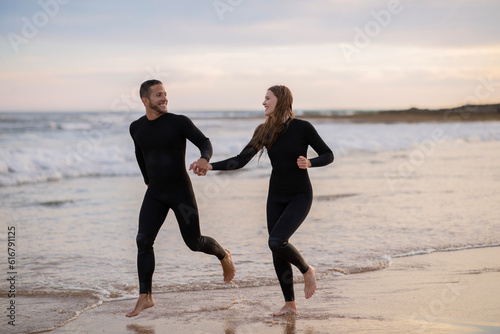Cheerful Young Couple Wearing Wetsuits Running On Seashore Together