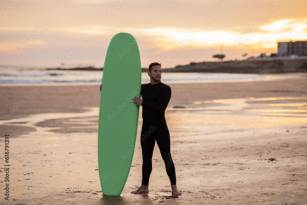 Handsome Surfer Man With His Surfboard Standing On The Beach At Sunset
