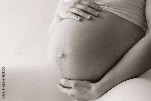 Midsection of a woman sitting and gently holding her very round pregnant baby bump. Side view. White background. Bright shot. Sepia.