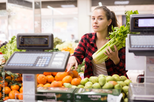 Billede på lærred Young female customer holding celery is about to buy tangerines, picking them ou