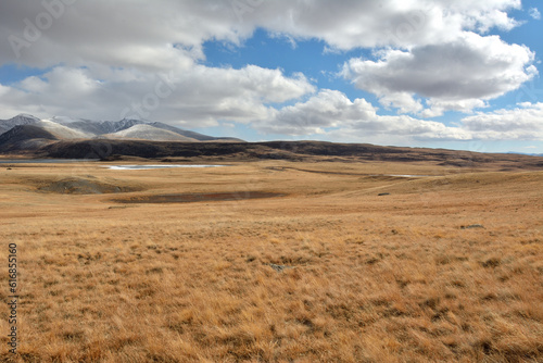 A huge flat steppe with dry  yellowed grass at the foot of snow-covered mountain ranges on a sunny autumn day.