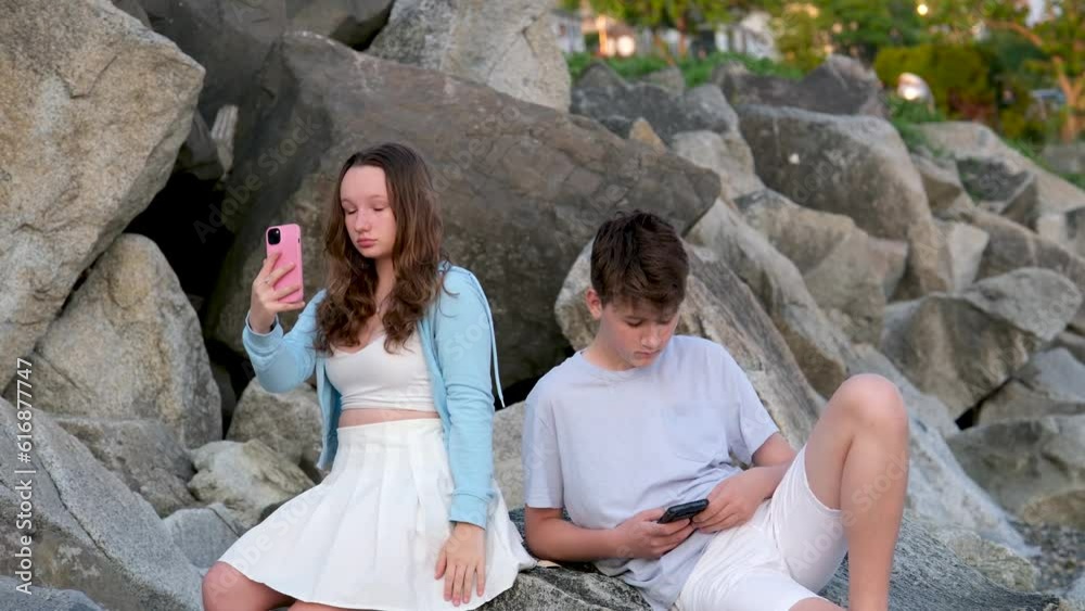 teens sitting on stones a boy and a girl with a phone in their hands use gadgets in nature dependent on social networks boredom on first date failed communication light clothes adolescence