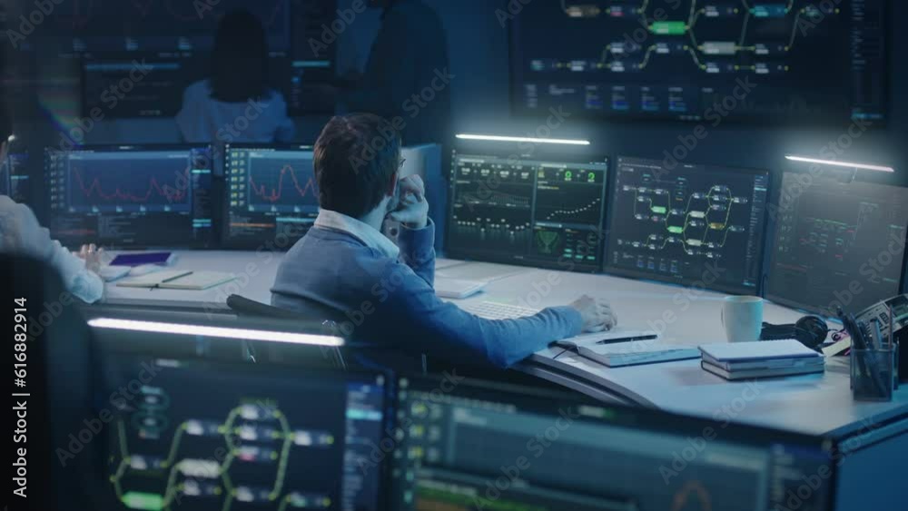 Software engineer works at computer in monitoring room late night. Big data scientists analyze server data. Multiple big screens on the wall with displayed real-time analysis charts. Cyber security.