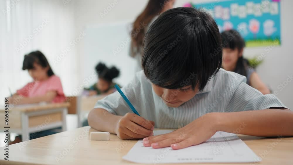 Diverse primary students doing an exam at elementary school. Teacher woman walking supervising examination of students at elementary school Diverse primary students doing an exam at elementary school. Teacher woman walking supervising examination of students at elementary school