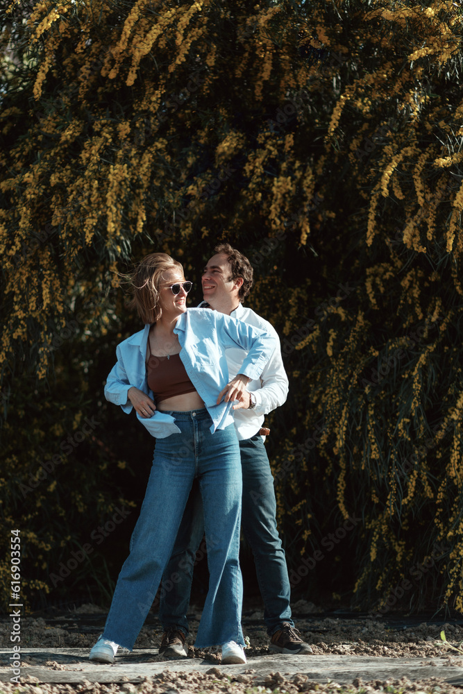Obraz premium Outdoor shot of young couple in love walking on pathway through field. Man and woman walking along yellow flowering trees.