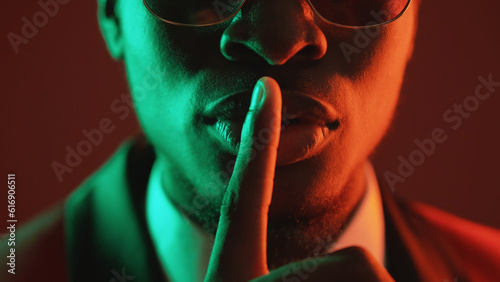 Shush gesture. Man secret. Confidential conspiracy. Green red neon light unrecognizable gentleman in suit glasses warning shh with finger on lips on dark.