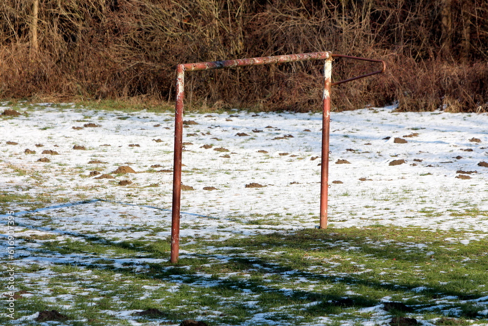Fototapeta premium Rusted old goal post without net left on abandoned children playground covered with freshly fallen snow mixed with uncut grass filled with multiple molehills next to dense forest vegetation
