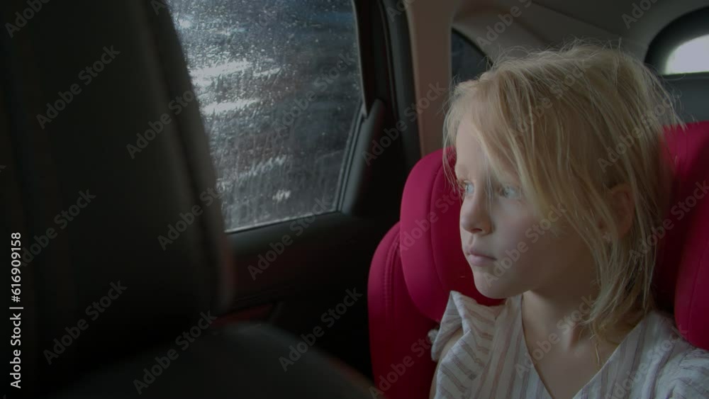 The little girl is sitting in the car's cabin, in a red child seat, and she is looking through the window as big brushes wash the car The little girl is sitting in the car's cabin, in a red child seat, and she is looking through the window as big brushes wash the car