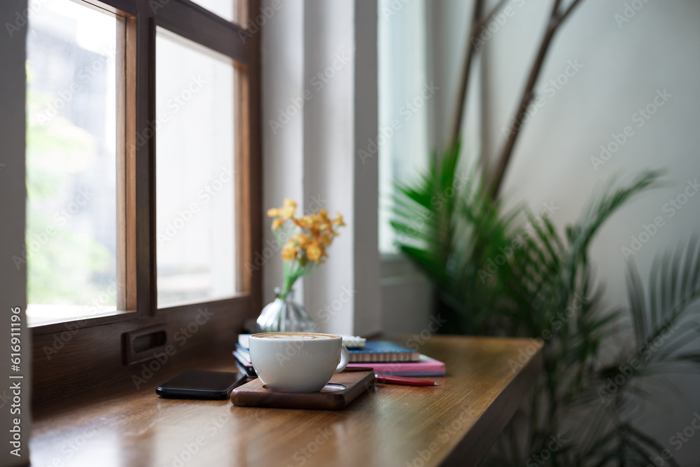 Fototapeta premium Close-up view, white coffee cup with latte coffee put on wooden counter and smart phone, note book put on digital tablet by window, coffee break and relax after work in cafe