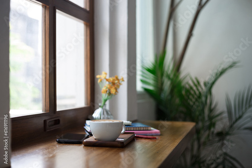 Close-up view, white coffee cup with latte coffee put on wooden counter and smart phone, note book put on digital tablet by window, coffee break and relax after work in cafe