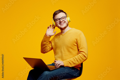 Excited man wearing yellow sweater and eyeglasses, using laptop computer while listening music in headphones, sitting on chair and looking away