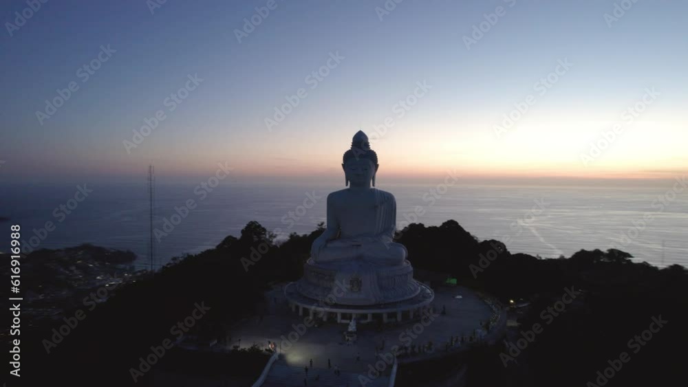 Big Buddha at sunset view from a drone. Phuket. People climb the steps to the statue. The green hills of the island are all around. In the distance, the bright sun goes over the sea. View from above Big Buddha at sunset view from a drone. Phuket. People climb the steps to the statue. The green hills of the island are all around. In the distance, the bright sun goes over the sea. View from above