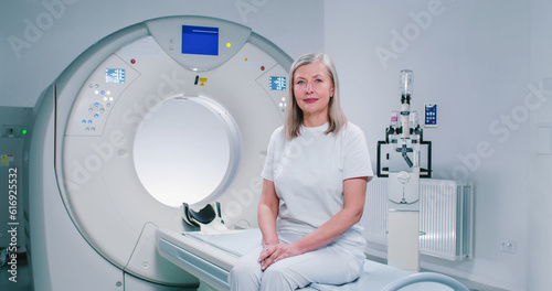 Shooting with smooth zoom. Mature woman sits at TC scanner bed. Female at room of MRI. Woman dressed up in white is looking at camera. Female patient is posing at background of medical equipment. photo
