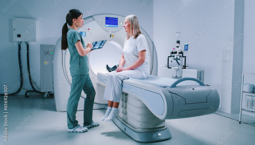 Doctor and patient are discussing results of tomography scanning. Female doctor is holding tablet and zooming scans of brain. Woman is sitting at CT scanner bed and listens attentively to doctor. Doctor and patient are discussing results of tomography scanning. Female doctor is holding tablet and zooming scans of brain. Woman is sitting at CT scanner bed and listens attentively to doctor.