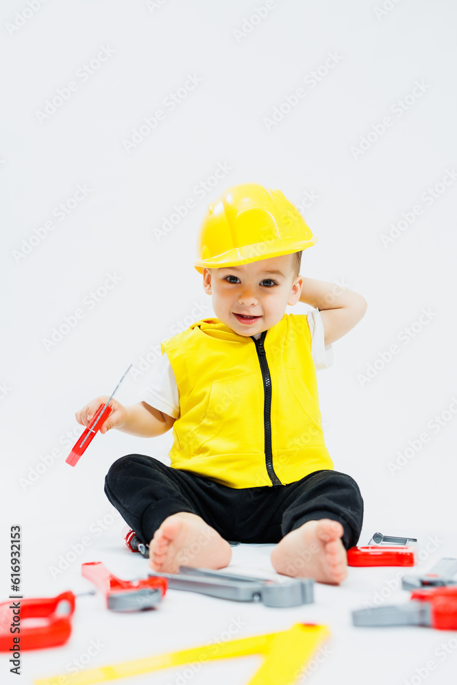 A two-year-old boy in a yellow vest sits and plays with plastic construction tools on a white background. Toys for small children. A two-year-old boy in a yellow vest sits and plays with plastic construction tools on a white background. Toys for small children.