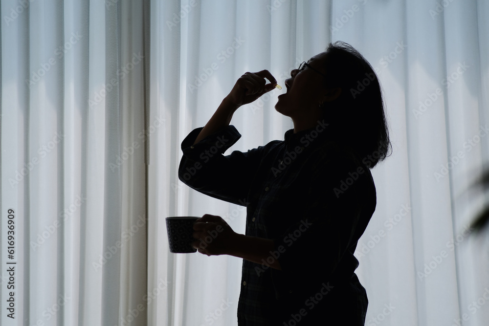 Silhouette of a woman standing by a window while taking a vitamin